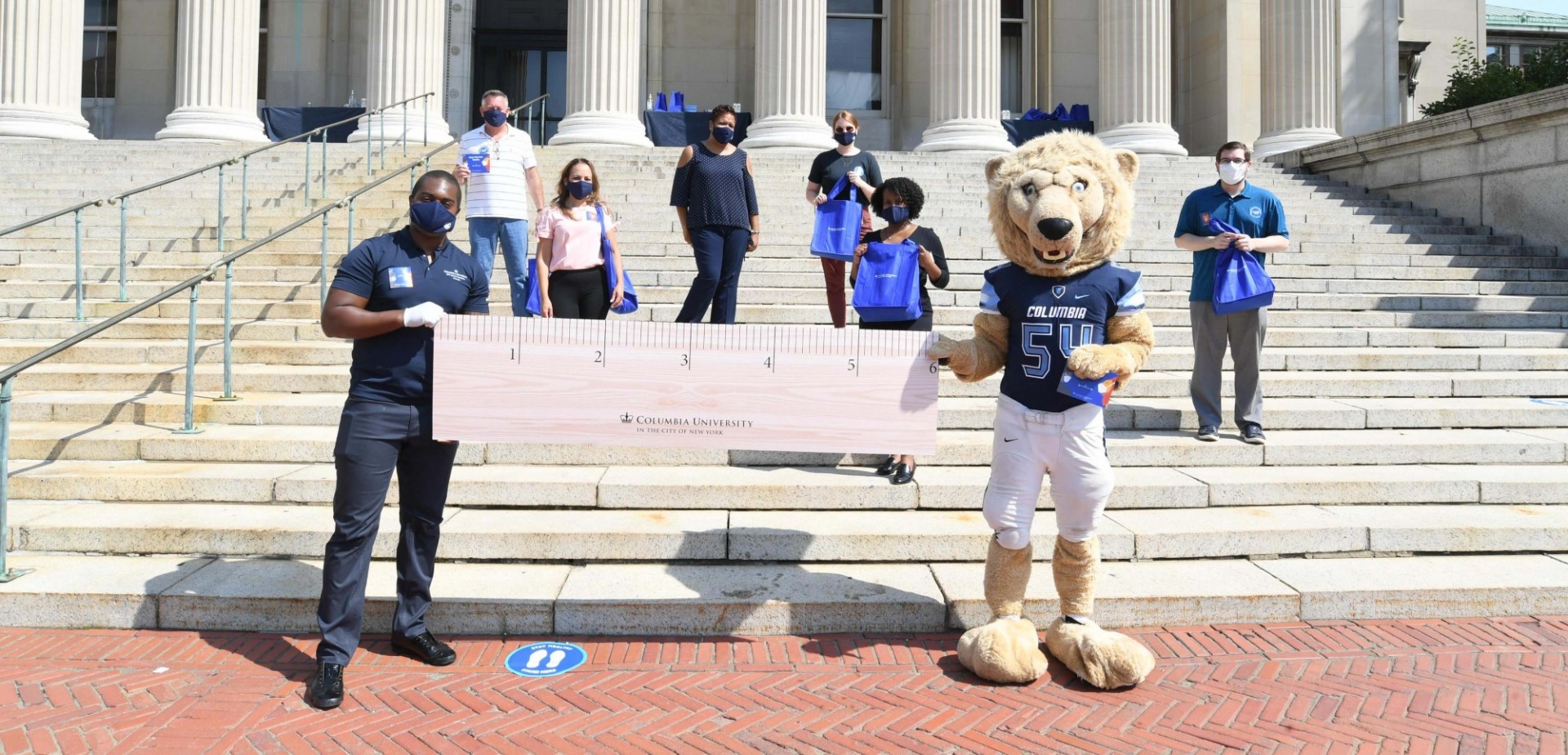 Facilities and Operations employees stand on the steps at Low Library holding a large six foot ruler with Roar-ee to demonstrate physical distancing.